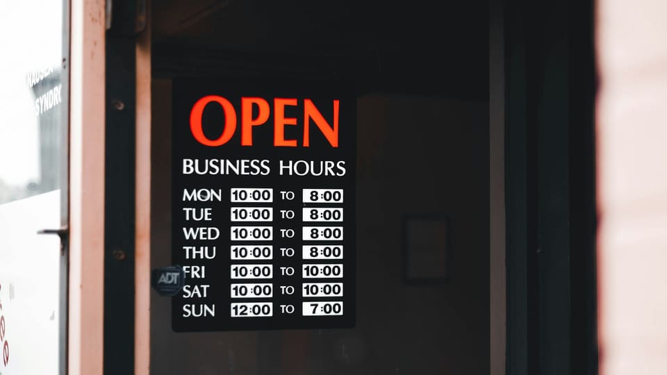 An OPEN sign with the business hours listed for each day, in a shop window