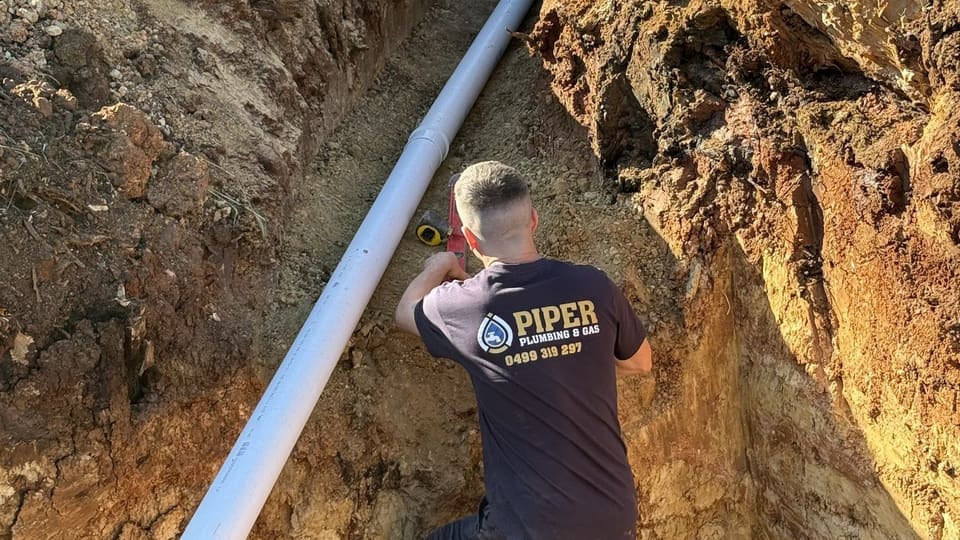 A Piper Plumbing & Gas plumber laying sewer pipe in a deep trench